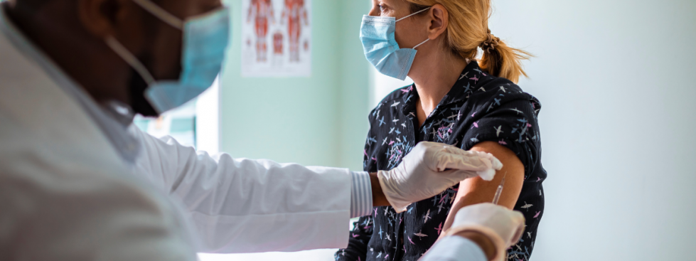 Male health care worker, wearing a lab coat, gives a female health care worker a vaccination in her arm.